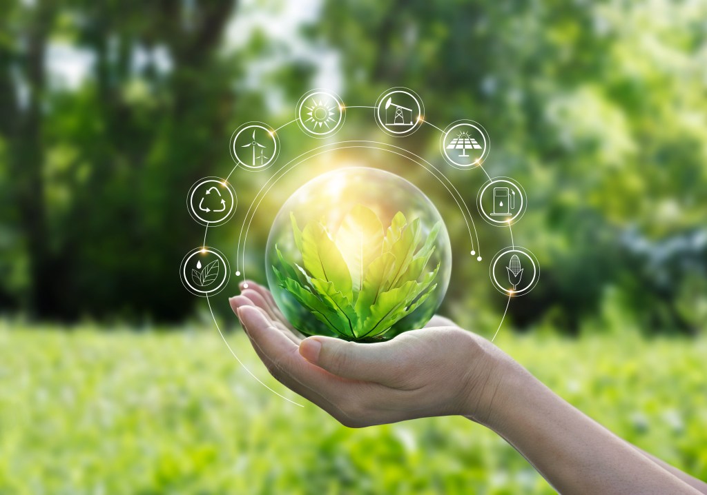 Photoillustration showing a hand holding and protecting a green growing plant in a bubble, in a green field surrounded by trees